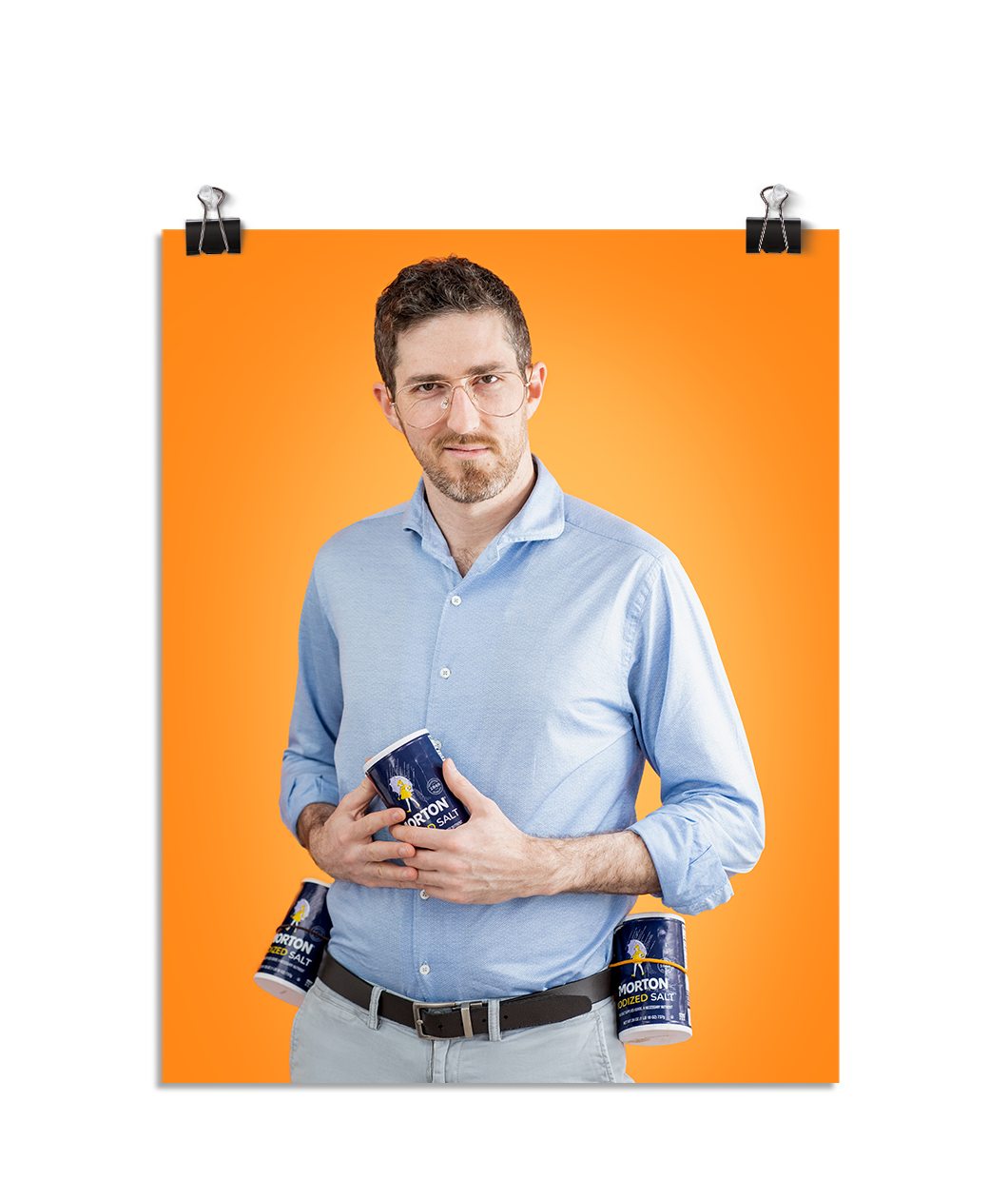 Photo of a man smirking at the camera with Morton's salt containers strapped to his belt on either side. He holds a third container of Morton's salt in his hands at his stomach. On an orange background.