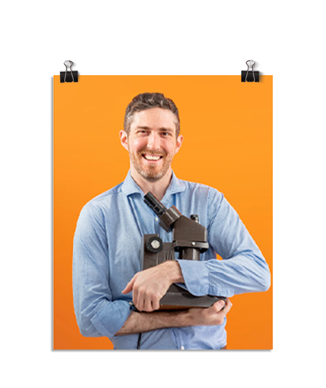 Photo of a man with a large smile wearing a light blue dress shirt as he holds his microscope to his chest like a proud father. On an orange background.