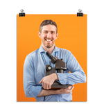 Photo of a man with a large smile wearing a light blue dress shirt as he holds his microscope to his chest like a proud father. On an orange background.