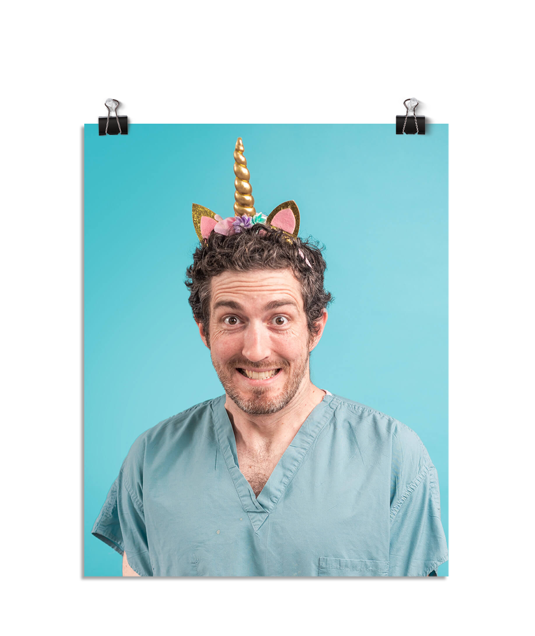 Photo of a man with an almost manic looking smile as he wears seafoam colored scrubs and a glittering gold unicorn horn headband on his head. On a seafoam colored background.