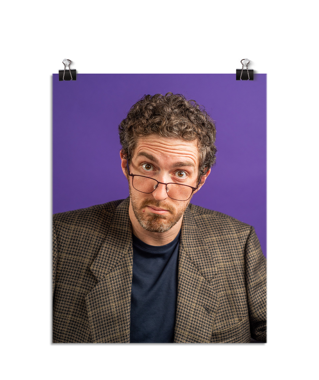 Photo of a man with his glasses notched low on his nose looking at the viewer with raised eyebrows. He is wearing a tweed suit jacket with a navy t shirt underneath and you can tell you did something to upset him. On a purple background.