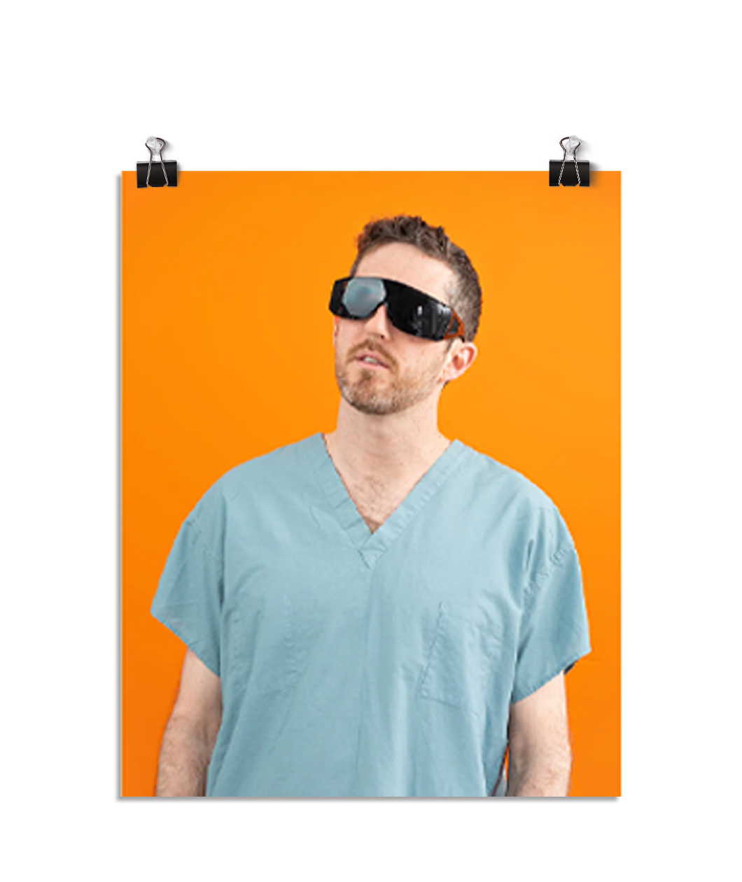 Photo of a man wearing large black glasses and seafoam colored scrubs looking up to the left. On an orange background.