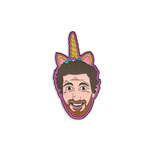 Enamel pin with black metal plating that has the head of a man smiling wearing a unicorn horn with glitter on it on a pink background.