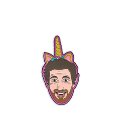 Enamel pin with black metal plating that has the head of a man smiling wearing a unicorn horn with glitter on it on a pink background.