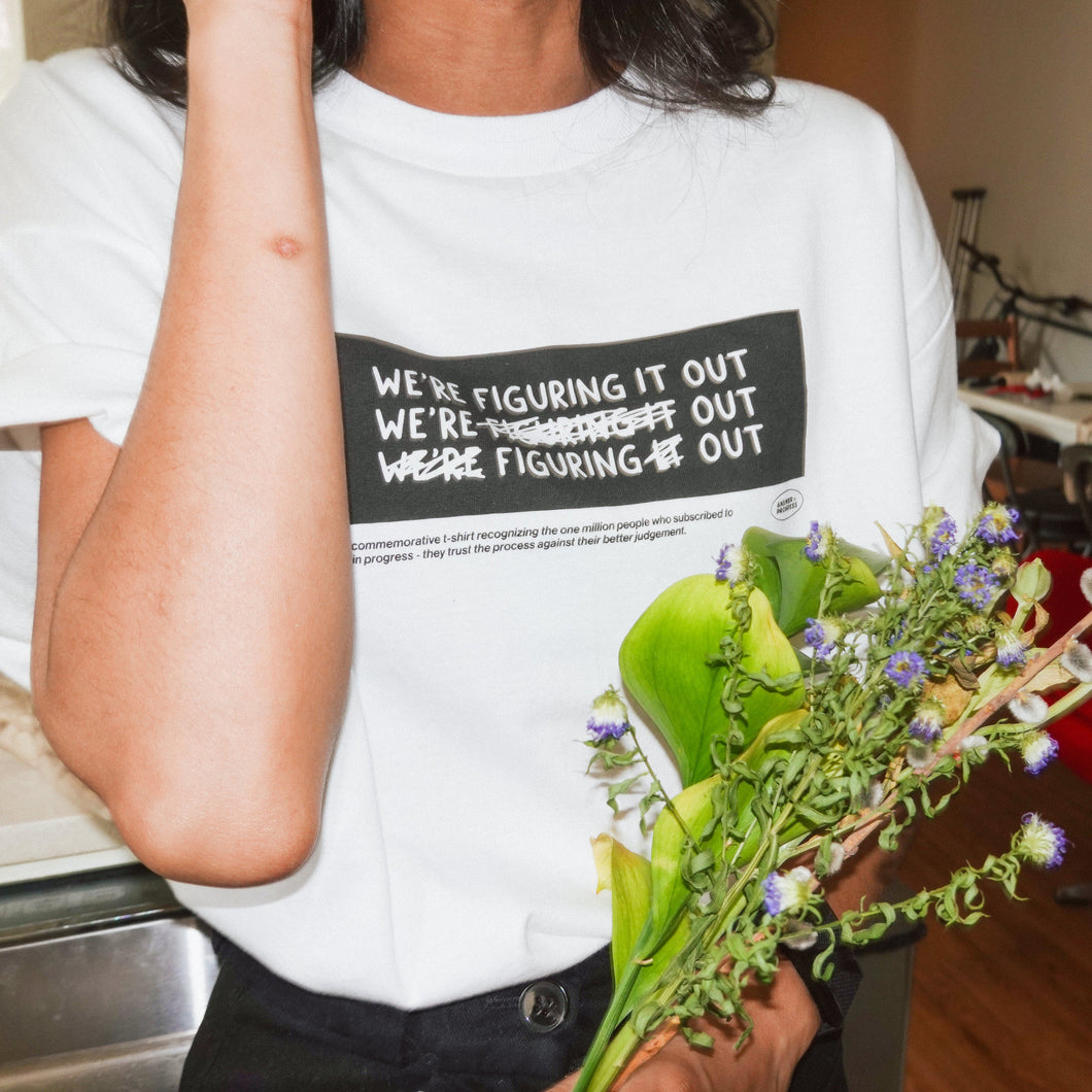 A photo of person's torso wearing a white t-shirt with a black rectangle in the center with the words "We're figuring it out" written three times in a row with the second line has "figuring" crossed out and the third line has "we're" and "it" crossed out. There is additional small text below this. From Answer in Progress.