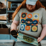 Person wearing a t-shirt with multiple logos, holding vinyl records in a storage room.