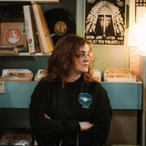 Woman wearing a black long sleeve crewneck crossing her arms in a record store. Crewneck has a teal record printed on the heard with an orange middle and says Hard Pivot back to physical media in the center of it with the DFTBA Records logo beneath.