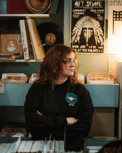 Woman wearing a black long sleeve crewneck crossing her arms in a record store. Crewneck has a teal record printed on the heard with an orange middle and says Hard Pivot back to physical media in the center of it with the DFTBA Records logo beneath.