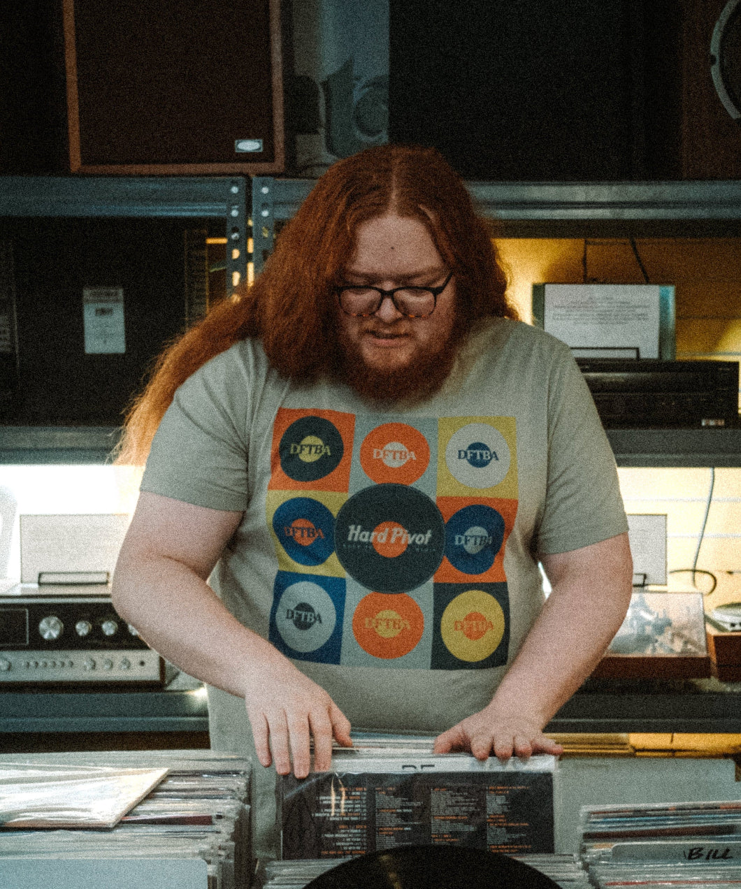 Man wearing a light green shirt with andy warhol style orange, teal, blue, and lime green 9 box print of records that say DFTBA in the middle of them. The center of the shirt has a teal record that says Hard Pivot back to physical media.