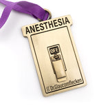 Gold metal rectangular ornament that says Anesthesia on the top, recessed into the metal, and then a 3D switch can flip up or down to indicate ON or OFF in the middle. The bottom has Dr.Glaucomflecken recessed into the metal. A ribbed purple ribbon is attached at the top so it can hang on a tree.