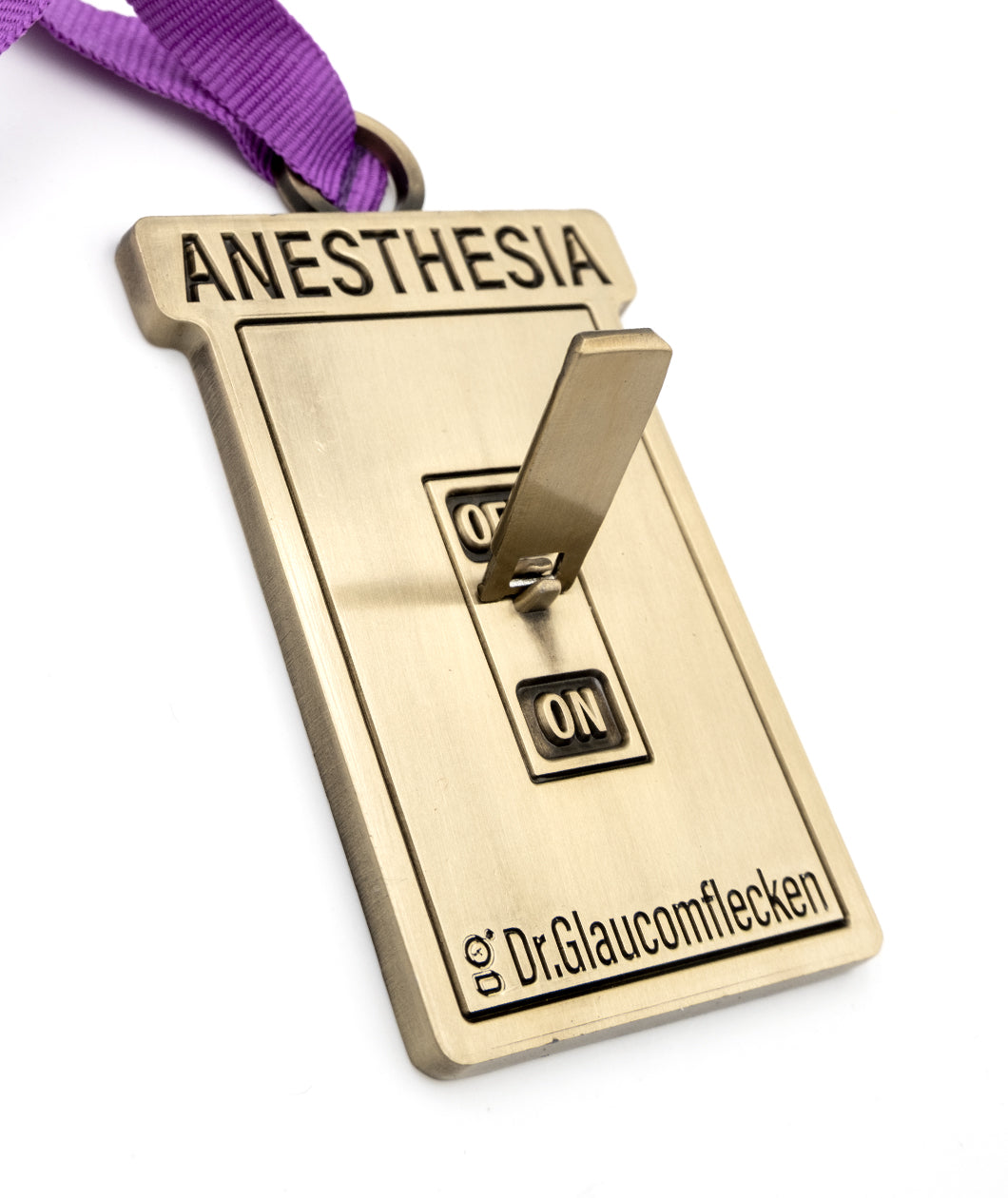 Gold metal rectangular ornament that says Anesthesia on the top, recessed into the metal, and then a 3D switch can flip up or down to indicate ON or OFF in the middle. The bottom has Dr.Glaucomflecken recessed into the metal. A ribbed purple ribbon is attached at the top so it can hang on a tree.