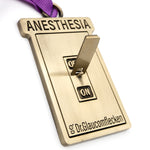 Gold metal rectangular ornament that says Anesthesia on the top, recessed into the metal, and then a 3D switch can flip up or down to indicate ON or OFF in the middle. The bottom has Dr.Glaucomflecken recessed into the metal. A ribbed purple ribbon is attached at the top so it can hang on a tree.