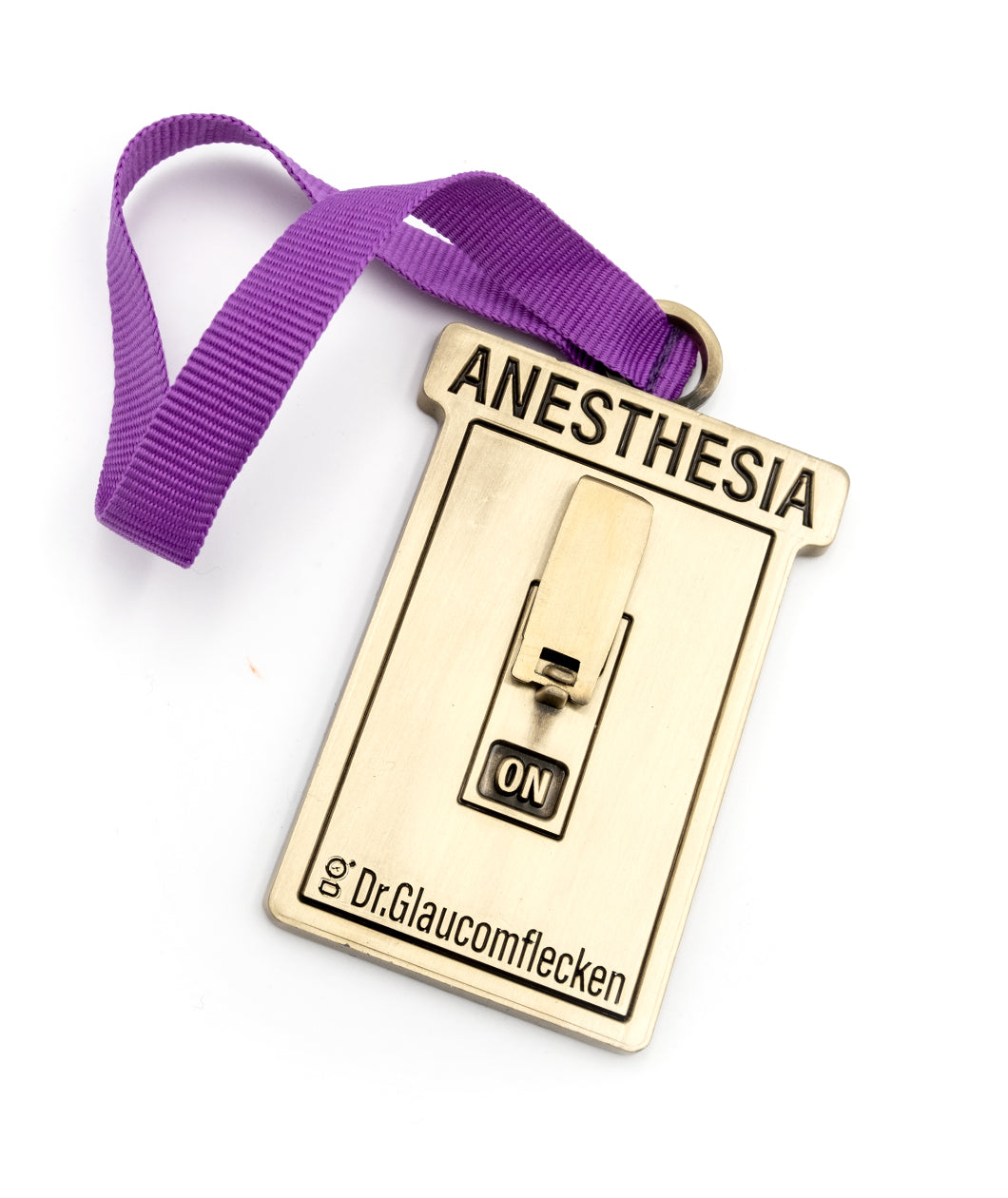 Gold metal rectangular ornament that says Anesthesia on the top, recessed into the metal, and then a 3D switch can flip up or down to indicate ON or OFF in the middle. The bottom has Dr.Glaucomflecken recessed into the metal. A ribbed purple ribbon is attached at the top so it can hang on a tree.