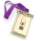 Gold metal rectangular ornament that says Anesthesia on the top, recessed into the metal, and then a 3D switch can flip up or down to indicate ON or OFF in the middle. The bottom has Dr.Glaucomflecken recessed into the metal. A ribbed purple ribbon is attached at the top so it can hang on a tree.