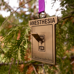 Gold metal rectangular ornament that says Anesthesia on the top, recessed into the metal, and then a 3D switch flipped up to indicate ON in the middle. The bottom has Dr.Glaucomflecken recessed into the metal. A ribbed purple ribbon is attached at the top that hangs it off of a Christmas tree.