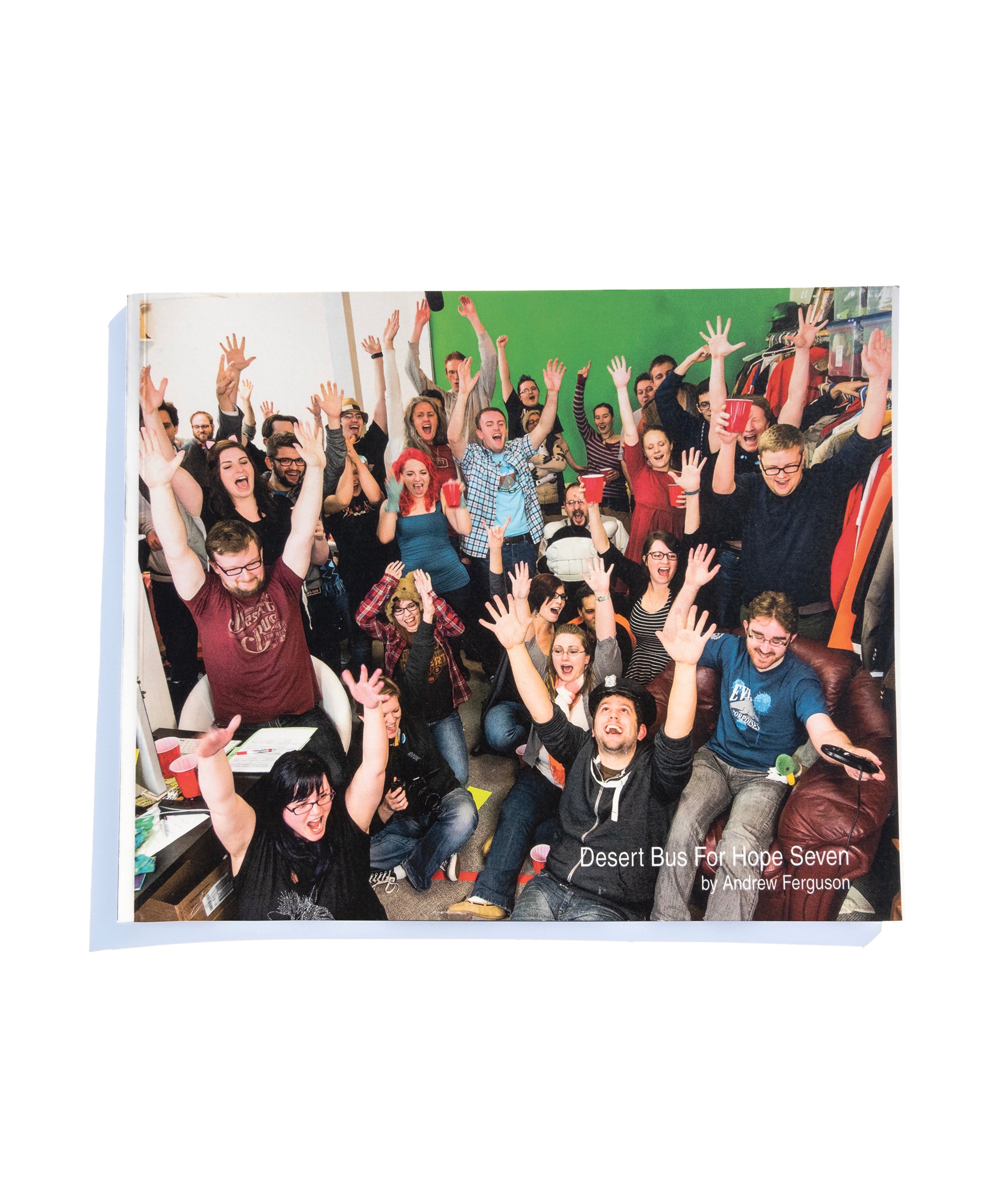 A photo of the front of the Desert Bus for Hope Seven photo book. It features a group of people from Desert Bus raising their hands in celebration.