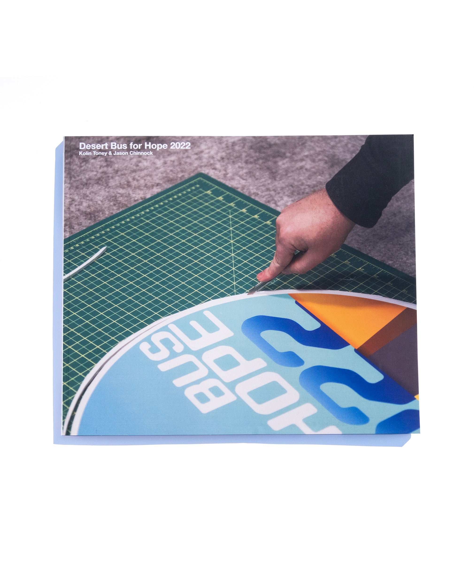 A photo of the front of the Desert Bus for Hope 2022 photo book. It features a hand trimming the margin off of a Desert Bus for Hope 2022 sign.