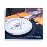 A photo of the front of the Desert Bus for Hope 2019 photo book. It features a paper plate that has a bus driving through the desert drawn on it.