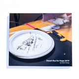 A photo of the front of the Desert Bus for Hope 2019 photo book. It features a paper plate that has a bus driving through the desert drawn on it.