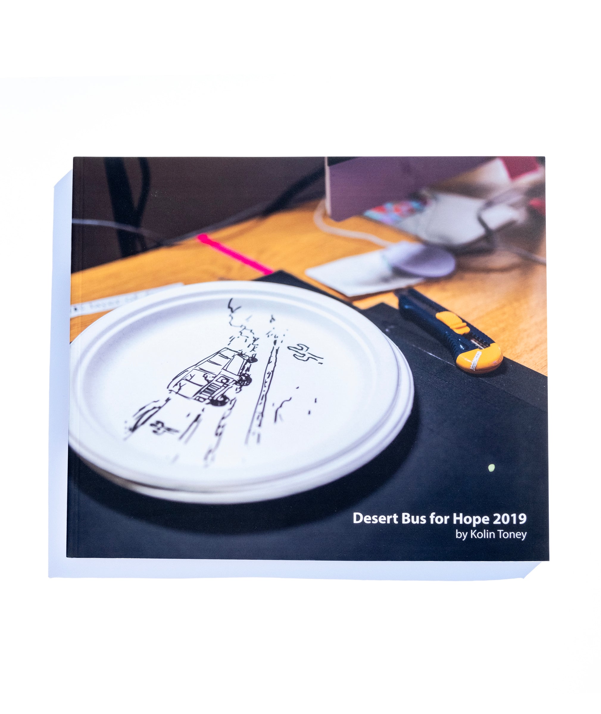 A photo of the front of the Desert Bus for Hope 2019 photo book. It features a paper plate that has a bus driving through the desert drawn on it.
