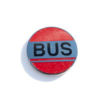 Photo of a round enamel pin with glittery read enamel stylized to look like a bus stop sign. It reads BUS in large black enamel letters