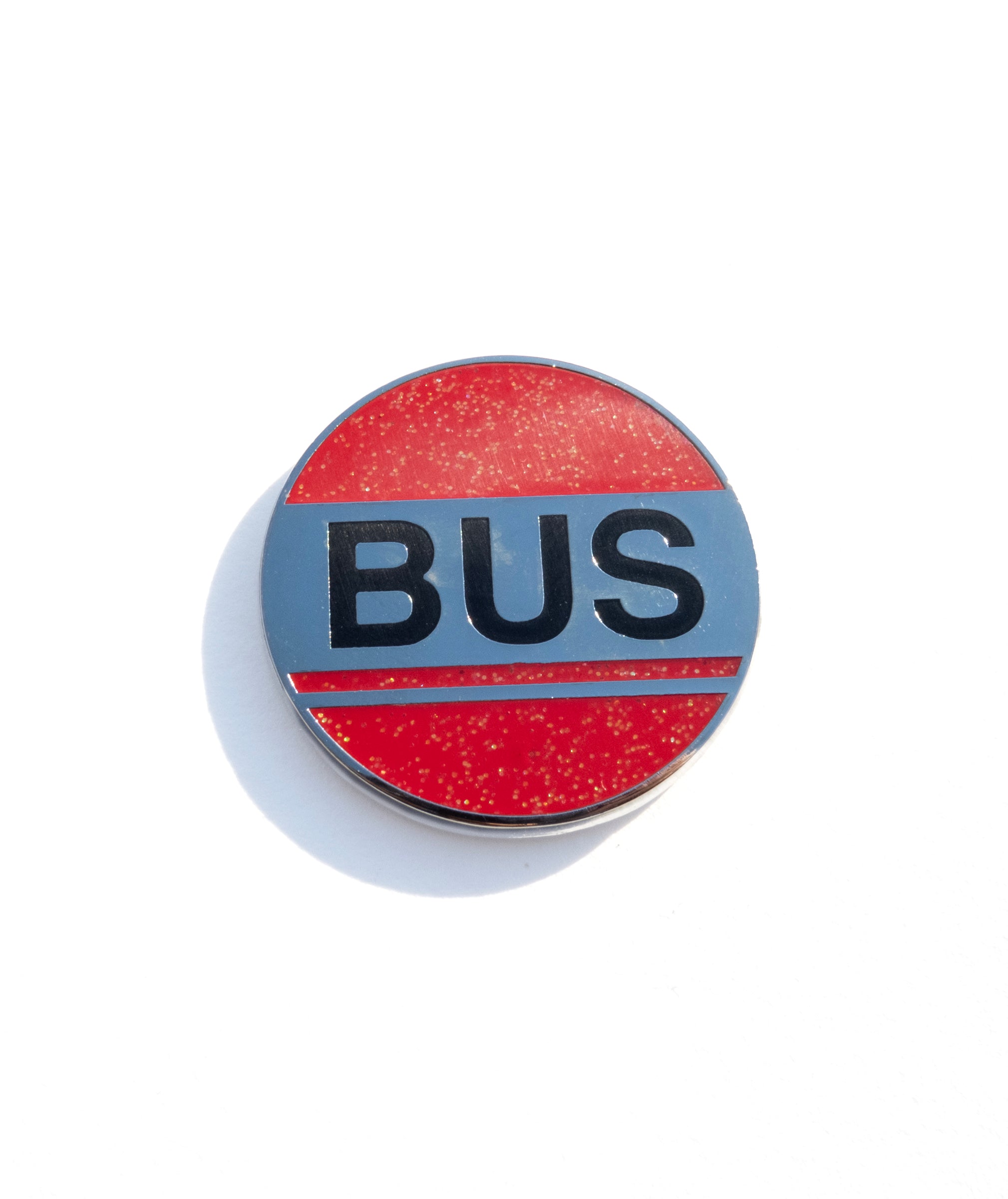 Photo of a round enamel pin with glittery read enamel stylized to look like a bus stop sign. It reads BUS in large black enamel letters