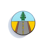 A photo of an enamel pin designed to show a road leading forward and a glittery tree-shaped air freshener above.