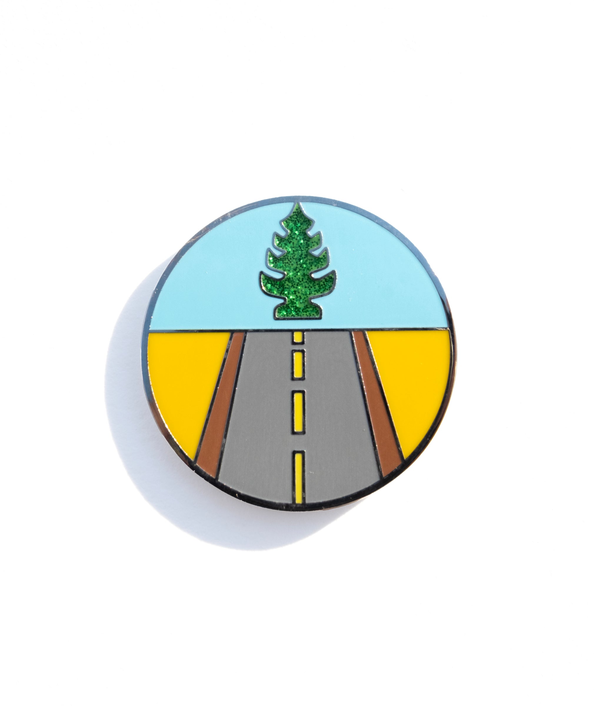 A photo of an enamel pin designed to show a road leading forward and a glittery tree-shaped air freshener above.