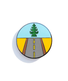 A photo of an enamel pin designed to show a road leading forward and a glittery tree-shaped air freshener above.