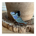 An enamel pin shaped like a blue bird wearing headphones, holding a cellphone that is playing TNO. 