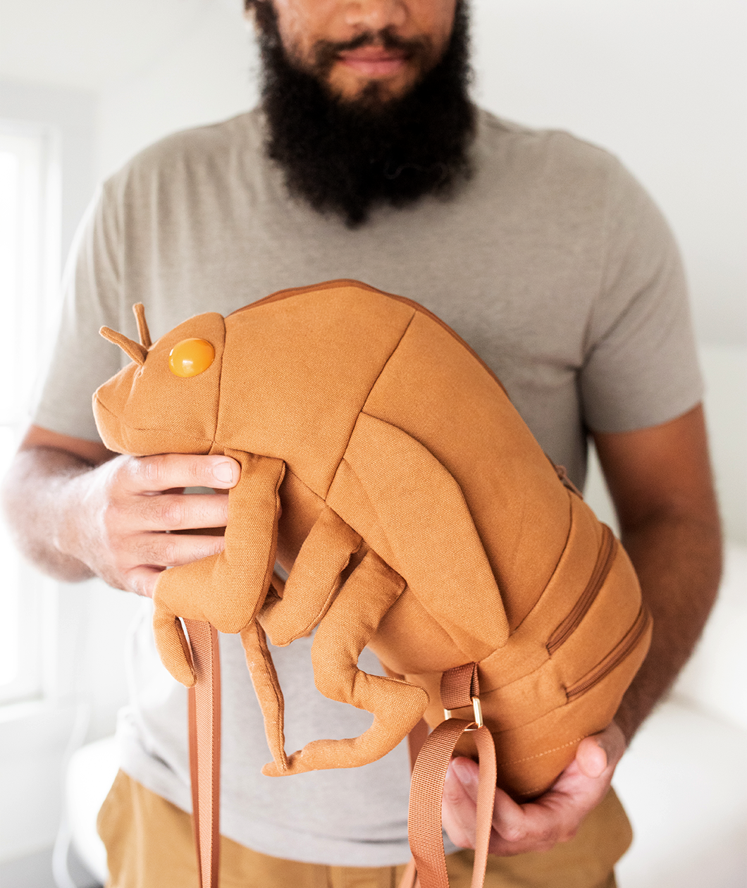 Man holding canvas backpack in the shape of a cicada shell complete with brown straps that attach the front legs to the thorax and egg yolk shaped protruding eyes.