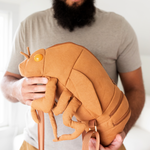 Man holding canvas backpack in the shape of a cicada shell complete with brown straps that attach the front legs to the thorax and egg yolk shaped protruding eyes.