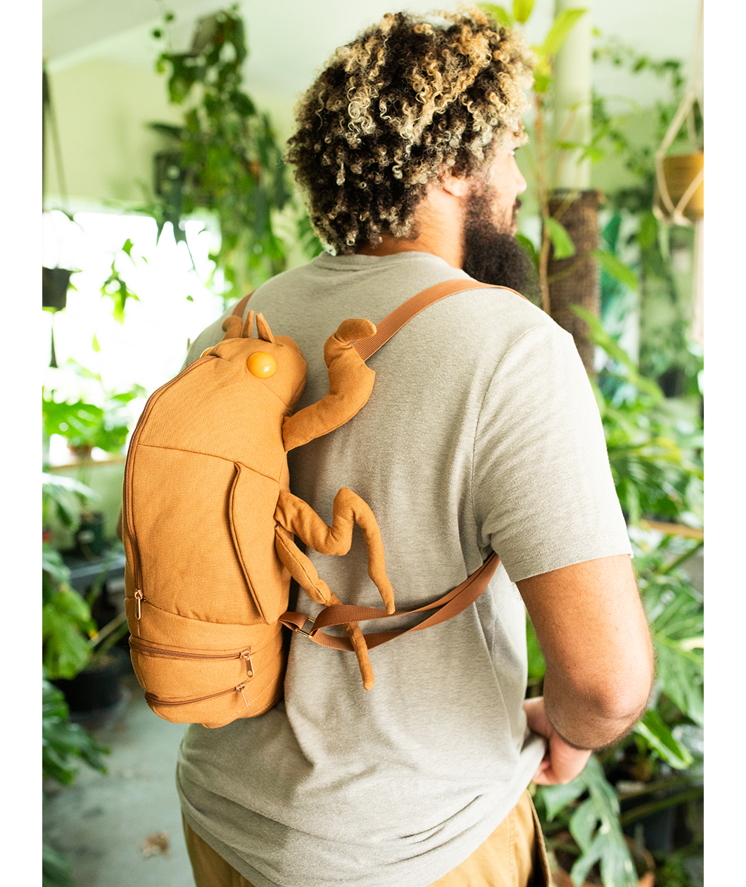 Man wearing cicada shell shaped backpack in a room full of plants. Looks like a giant cicada crawling up his back.