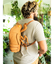 Man wearing cicada shell shaped backpack in a room full of plants. Looks like a giant cicada crawling up his back.