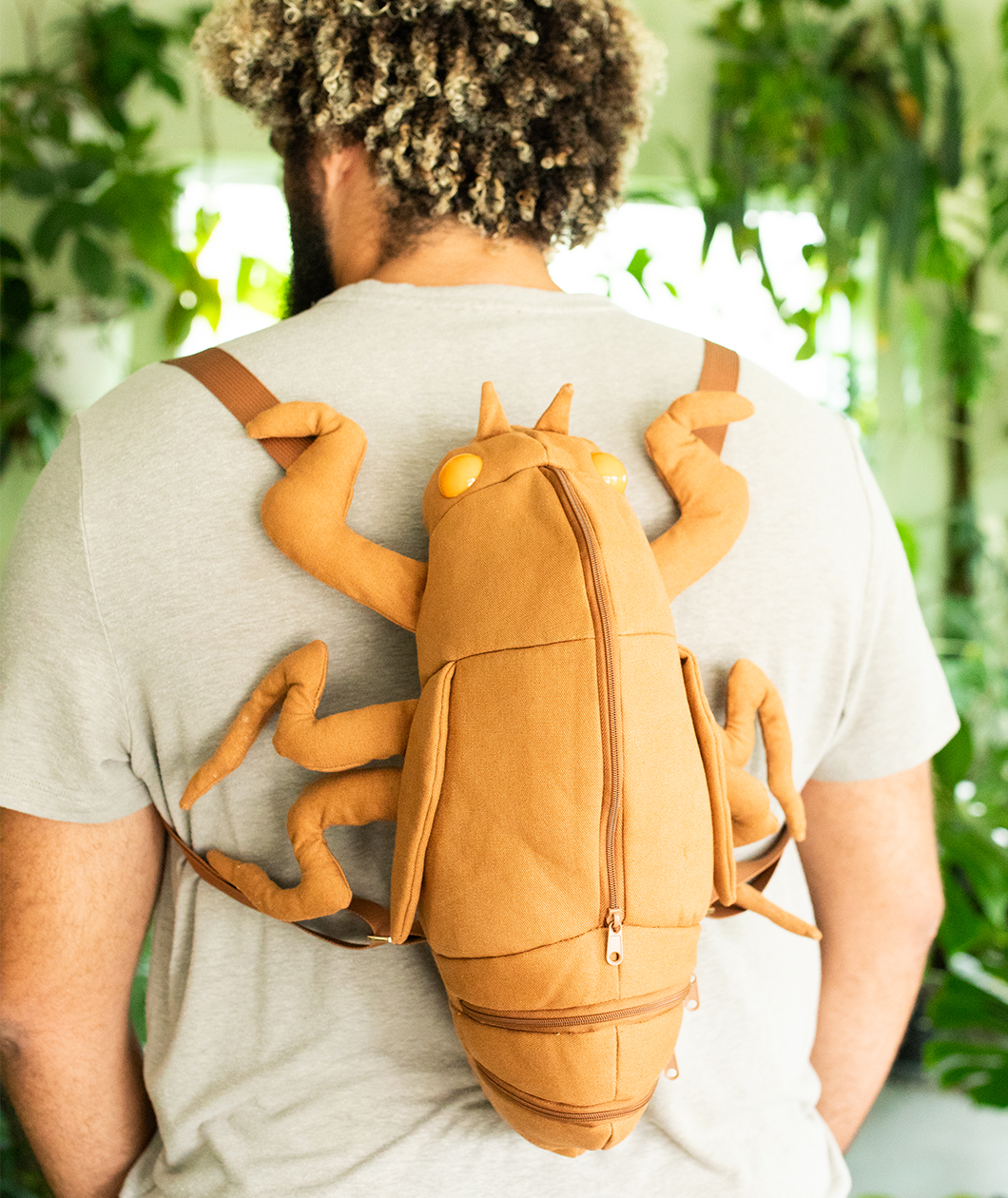 Man wearing cicada shell shaped backpack in very sunny lighting in a room with many plants.
