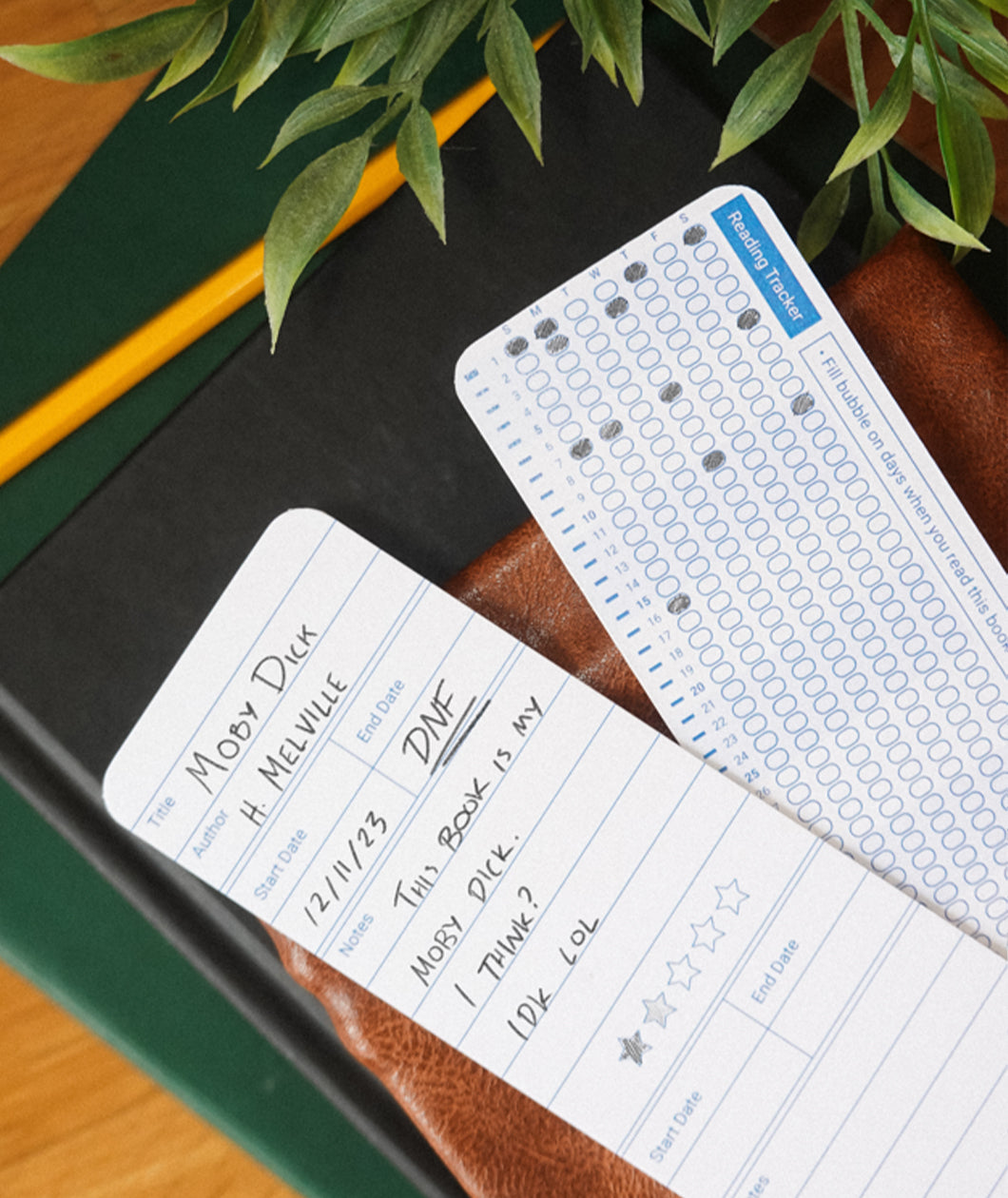 A set of 2 bookmarks showing the front and back designs. The bookmarks lay atop of a stack of books on a desk. From Answer in Progress.