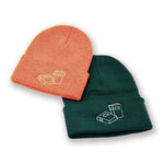 A pair of Books Unbound beanies, one of each color: Heather Orange and Forest. Each are embroidered with two books on the front of the cuff.