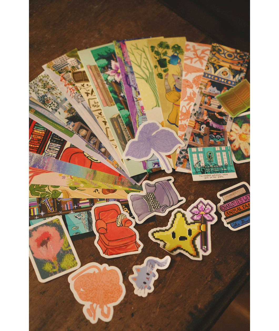 An assortment of differently designed paper bookmarks and die cut stickers spread out across a table top. From Books Unbound.