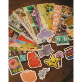 An assortment of differently designed paper bookmarks and die cut stickers spread out across a table top. From Books Unbound.