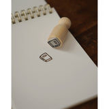 A photo of a miniature stamp of a book illustration. The stamp has been used on a fresh page of a notepad on a table. From Books Unbound.