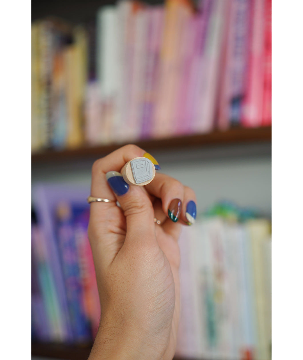 A photo of a miniature stamp of a book illustration. The stamp is being held up in front of a colorful bookcase. From Books Unbound.