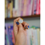 A photo of a miniature stamp of a book illustration. The stamp is being held up in front of a colorful bookcase. From Books Unbound.