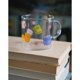 A glass mug with a design of colorful books printed all around. The mug sits atop a stack of books. From Books Unbound.