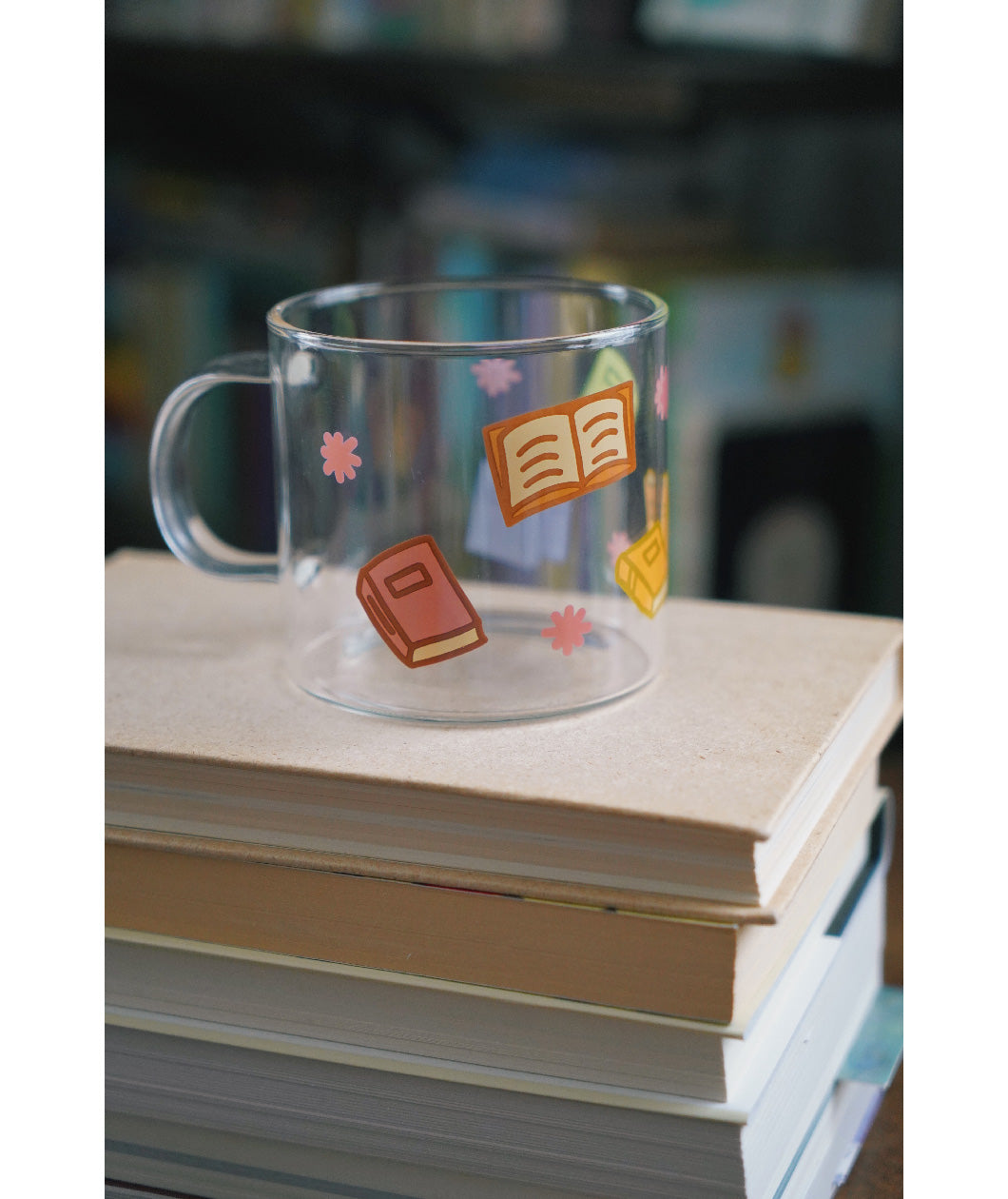 A glass mug with a design of colorful books printed all around. The mug sits atop a stack of books. From Books Unbound.