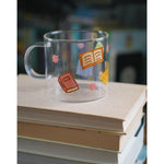 A glass mug with a design of colorful books printed all around. The mug sits atop a stack of books. From Books Unbound.