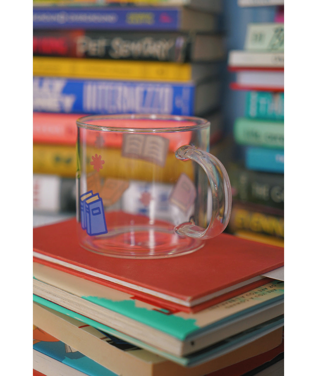 A glass mug with a design of colorful books printed all around. The mug sits atop a stack of books and is surrounded by other stacks of books. From Books Unbound.