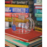 A glass mug with a design of colorful books printed all around. The mug sits atop a stack of books and is surrounded by other stacks of books. From Books Unbound.