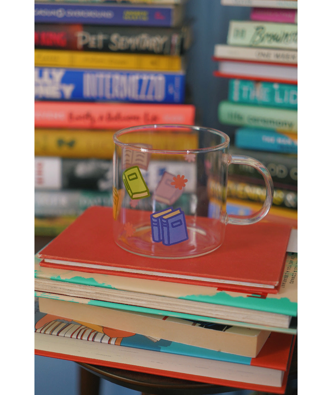 A glass mug with a design of colorful books printed all around. The mug sits atop a stack of books and is surrounded by other stacks of books. From Books Unbound.