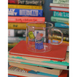 A glass mug with a design of colorful books printed all around. The mug sits atop a stack of books and is surrounded by other stacks of books. From Books Unbound.