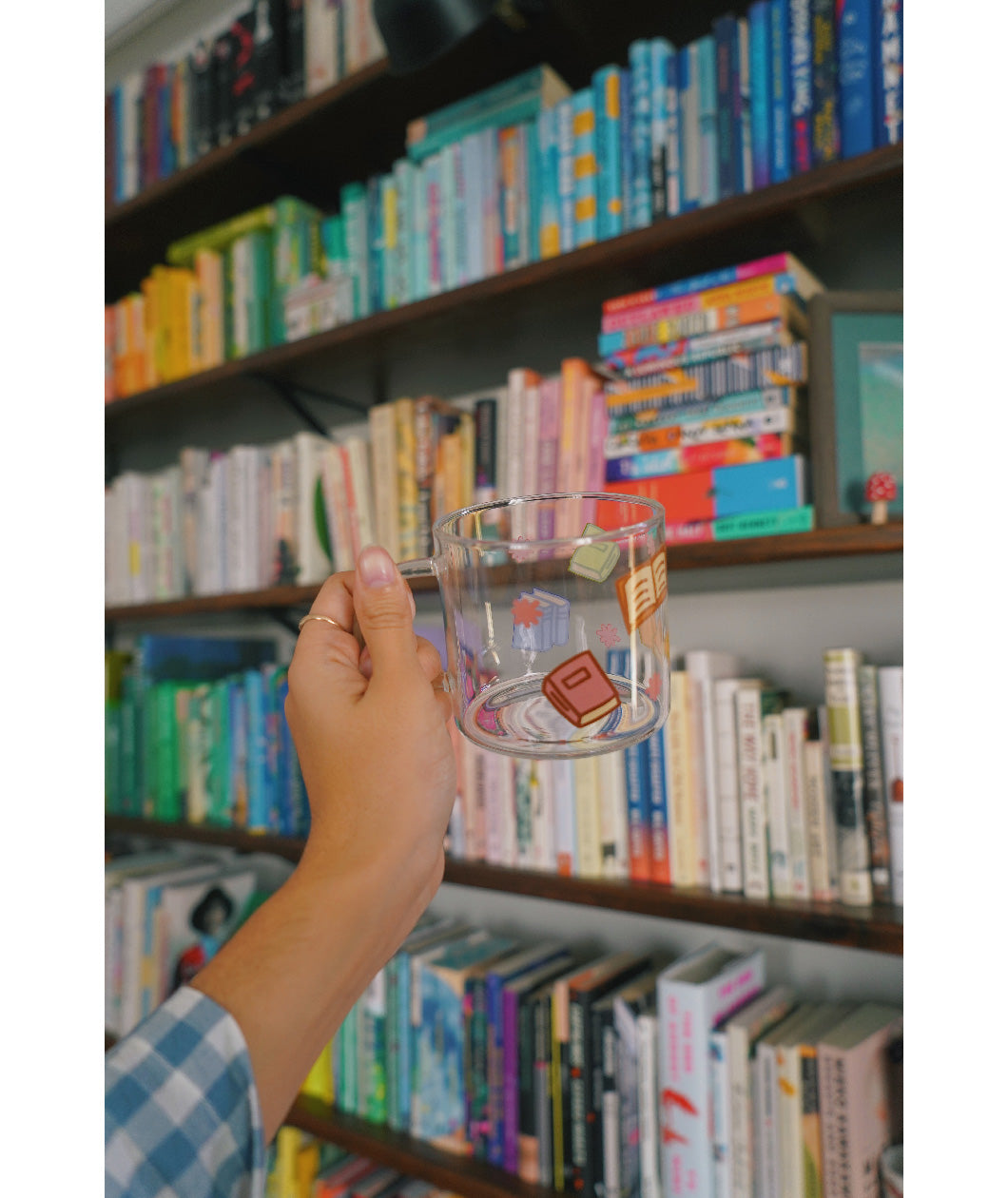 A glass mug with a design of colorful books printed all around. The mug is held up by a hand in front of a bookcase full of colorful books. From Books Unbound.